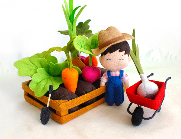 Felt hand sewn PDF tutorial with patterns by FeltCraftsFairyland - farmer baby boy in the blue overalls and a hat is standing near big vegetables in the beds in fencing, garden wheelbarrow and shovel