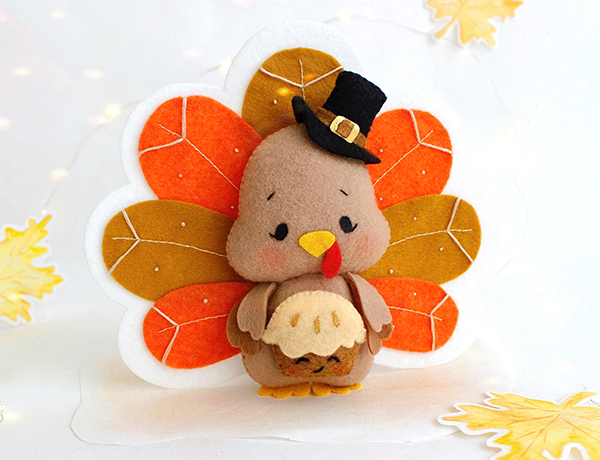 Felt hand sewn PDF tutorial with patterns by FeltCraftsFairyland - Thanksgiving turkey in black pilgrim hat with pumpkin pie in the wings is standing in front of the painted yellow maple leaves