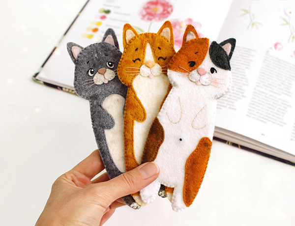 Felt hand sewn PDF tutorials with patterns by FeltCraftsFairyland - long gray, ginger and calico cats bookmarks in the authors hand in front of the opened book