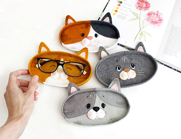 Felt hand sewn PDF tutorials with patterns by FeltCraftsFairyland - ginger, calico, tricolored and grey cats faces eyeglasses holders with authors hands near the opened book, the glasses laying on the ginger cat