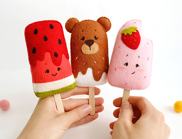 Felt hand sewn PDF tutorials with patterns by FeltCraftsFairyland - watermelon popsicle with wooden stick in the authors hand near the opened cook book, ice cream brown chocolate bear and pink strawberry popsicles