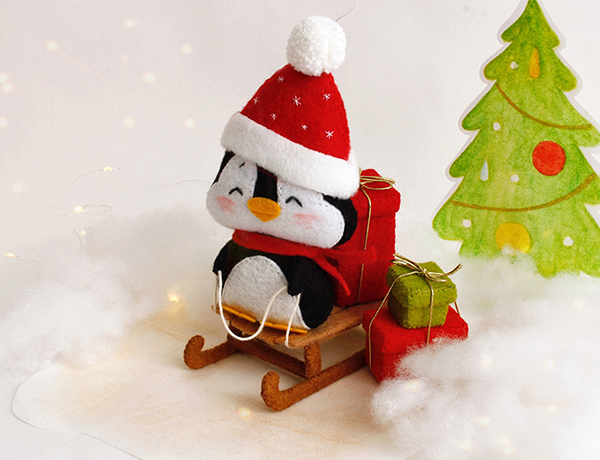 Felt PDF tutorial with patterns by FeltCraftsFairyland - hand sewn penguin in the red winter cap is seating on the sleigh with Christmas red and green box presents