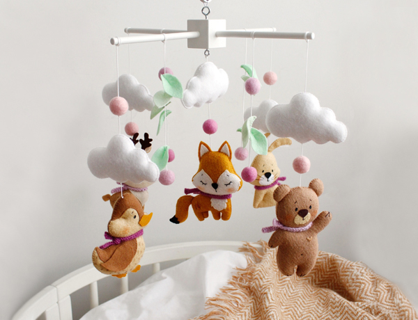 Felt hand sewn woodland baby crib mobile with light brown bear, duck, bunny, fox and deer, wool balls and many leaves is hanging over a baby cot for a newborn with cozy cotton blanket in it